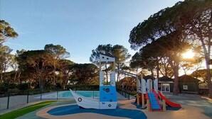 Ein Spielplatz mit Rutsche und Klettergerüsten, umgeben von Bäumen und einem eingezäunten Bereich.