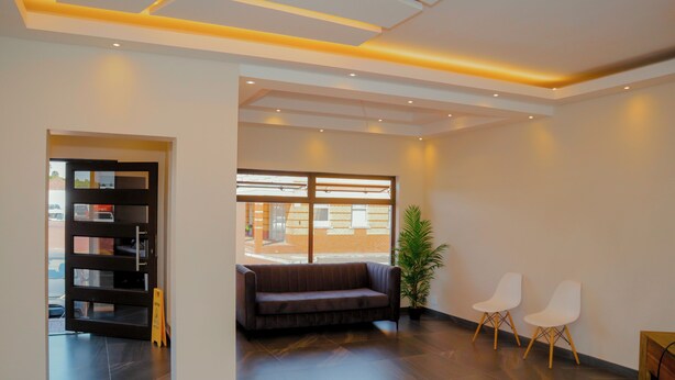 A modern interior with a dark wood floor, a brown sofa, two white chairs, a potted plant, and a bookshelf.