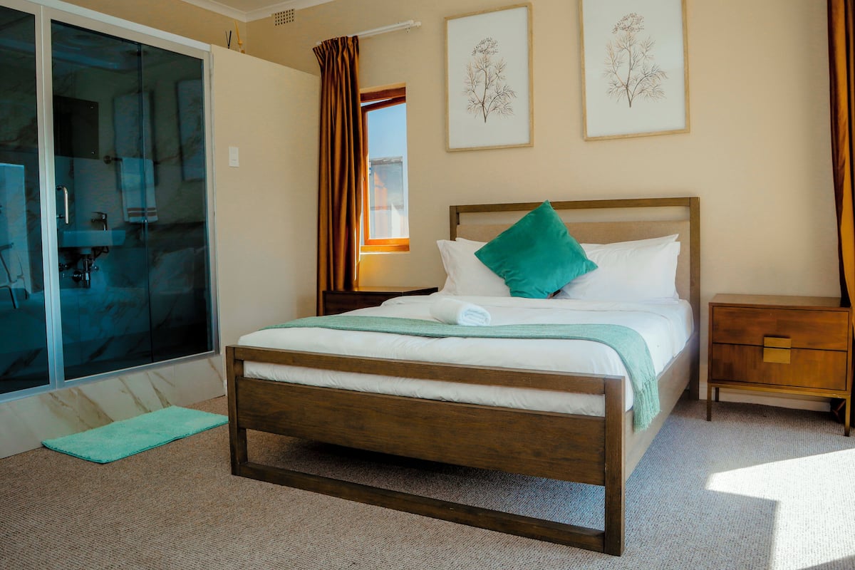 Quarto com cama de madeira, travesseiro verde, mesa de cabeceira e duas gravuras botânicas emolduradas na parede.