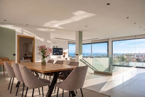 A modern dining area with a wooden table and chairs, a TV, and a view of the city and ocean.