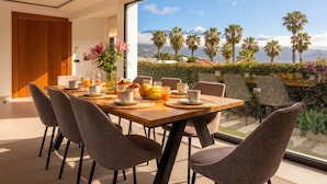 Un comedor con una mesa preparada para el desayuno, un amplio ventanal con vistas a palmeras y una montaña, y una decoración de estilo moderno.