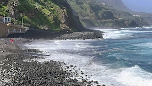 Una playa rocosa con olas, un edificio con balcón y una montaña al fondo.