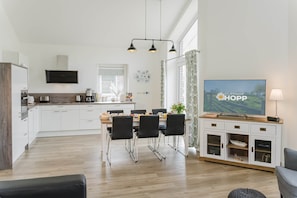 Une cuisine moderne avec des meubles blancs, un coin repas avec des chaises noires et un téléviseur diffusant l’émission « BAUERNHOF HOPF ».