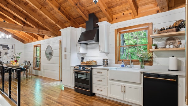 A kitchen with wooden beams, white cabinets, a black stove, and a window with a view of greenery.