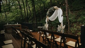 An outdoor venue with wooden chairs arranged in rows, a rustic wooden table, and a decorative arch with drapery and greenery.