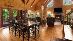 A wooden cabin interior with a dining area, fireplace, and large windows. - Secluded Creekside Cabin - Fire Pit, Deck & Mountain Views, Sleeps 8 (Lake Lure)