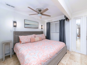 Relax in this serene bedroom with a large bed and ceiling fan, featuring a window with blinds and a door to a separate room.