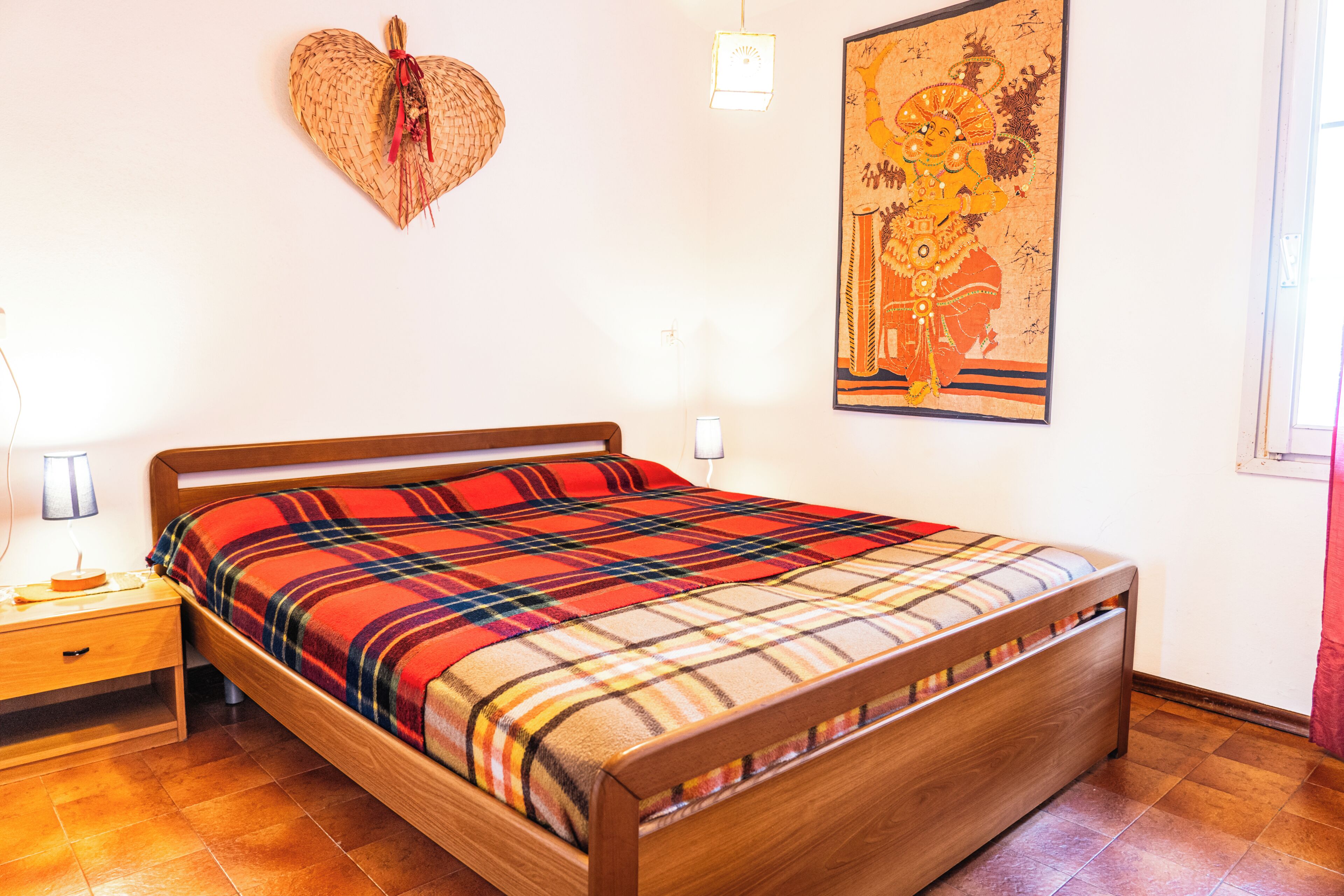 Uma cama de madeira com colcha xadrez, uma mesinha de cabeceira com abajur, uma decoração em formato de coração na parede e um quadro emoldurado.