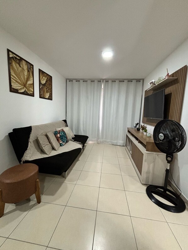 A living room with a sofa, a small table, a fan, and a TV mounted on the wall.