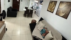 A living room with a black sofa, a coffee table, and a dining area with chairs and a table. There is a kitchen in the background with a refrigerator and a microwave.