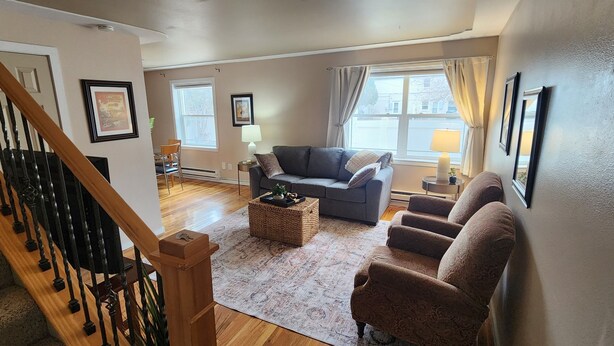 A spacious living room with a staircase, a grey sofa, a patterned rug, and a dining area with a table and chairs.