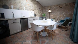 Una cucina compatta con parete in pietra, zona pranzo con tavolo rotondo e sedie, e angolo relax con poltrona e lampada da terra.