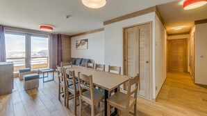 A modern room with a wooden dining table, chairs, and a sofa. There are large windows with curtains, a door, and a hallway.