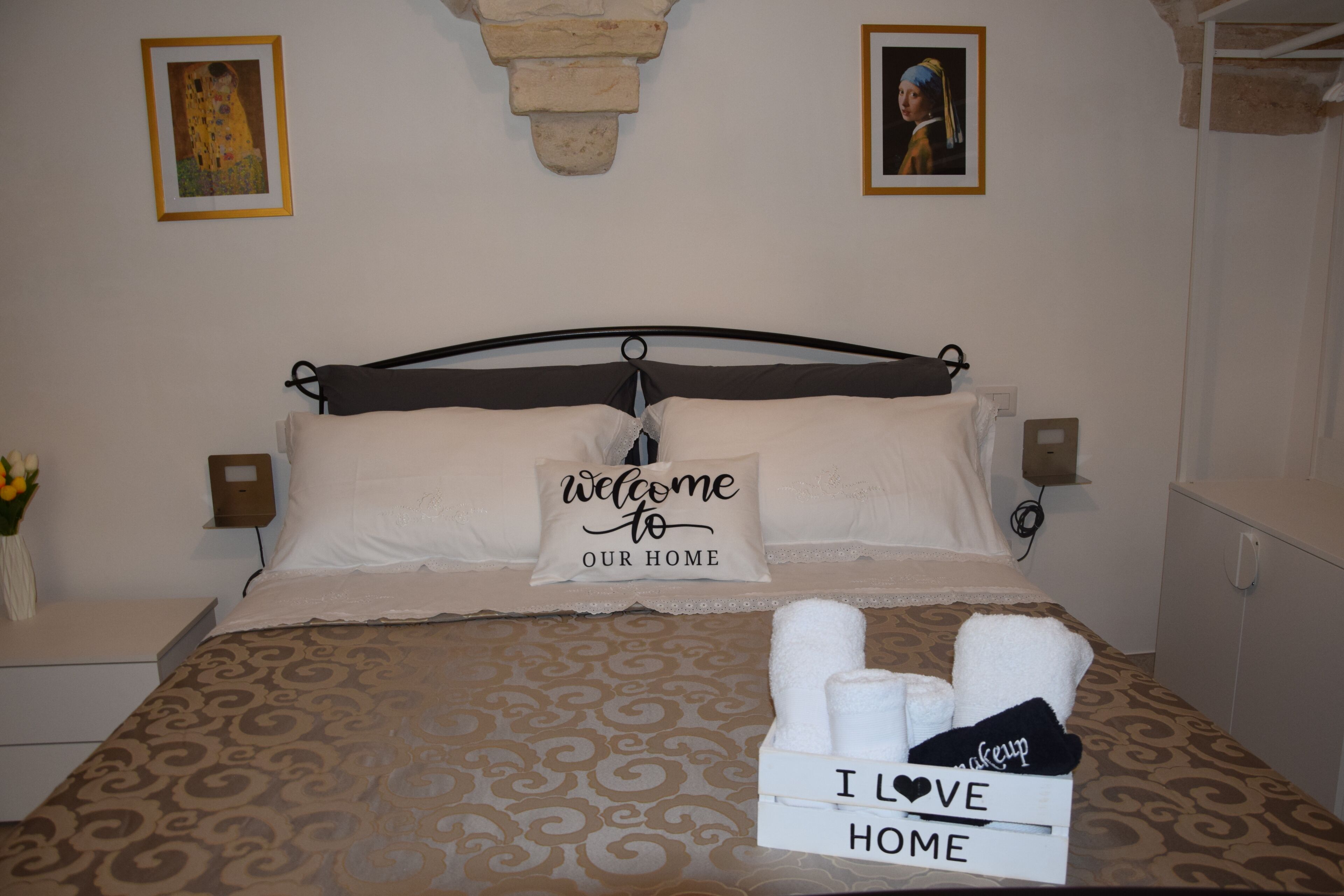 Ein ordentlich bezogenes Bett mit einem Kissen mit der Aufschrift 'Willkommen in unserem Zuhause', einer Box mit 'Ich liebe Zuhause' und zwei gerahmten Bildern an der Wand.