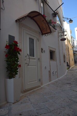 Une ruelle étroite pavée, bordée d’un bâtiment blanc qui présente une porte, une petite fenêtre et un balcon orné de plantes en pot.