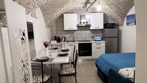 Cocina compacta con armarios blancos, un lavamanos y una estufa. Hay una zona de comedor con una mesa y sillas, y una cama con cubierta azul.