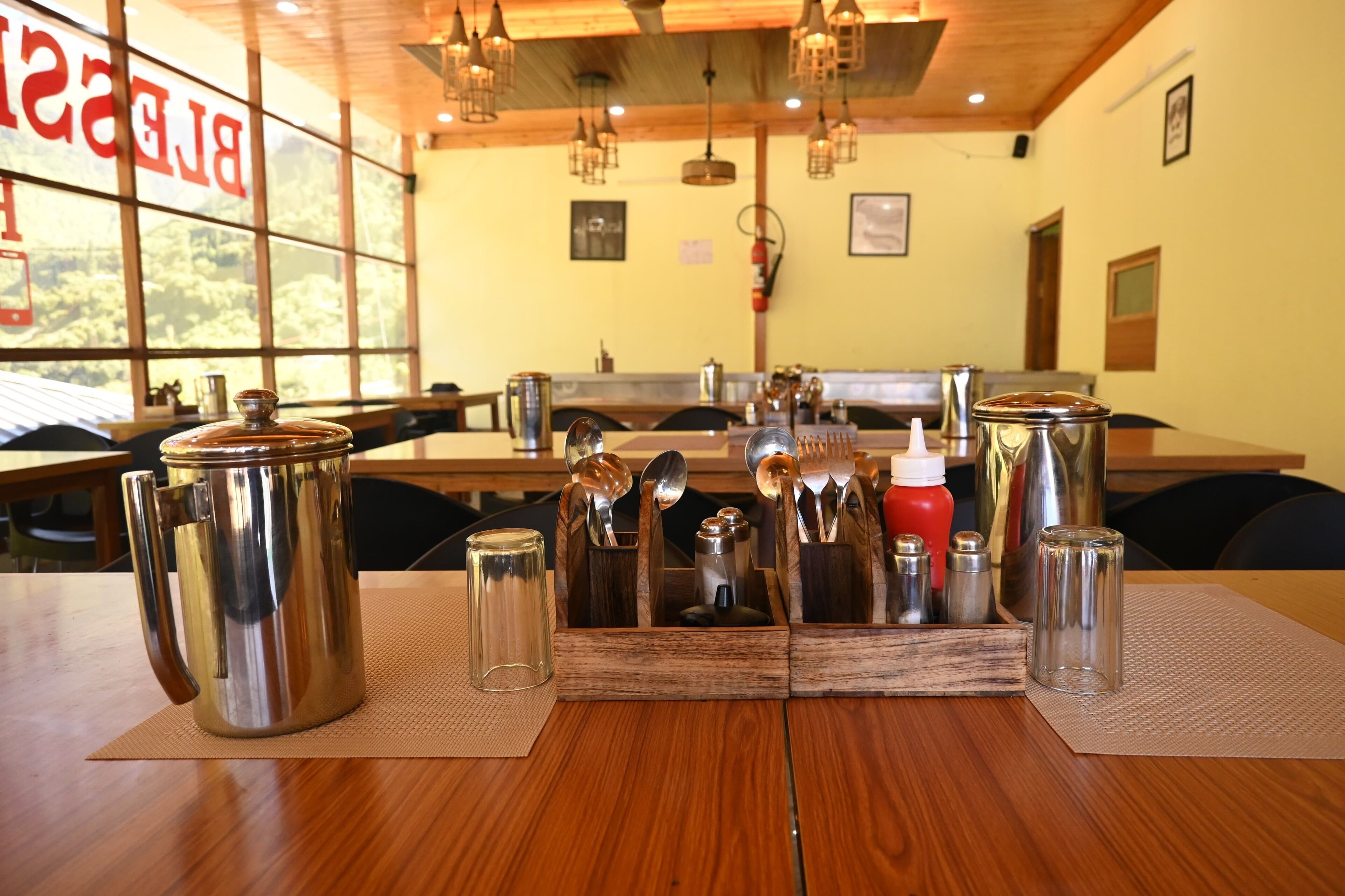 A restaurant with wooden tables, metal serving utensils, and a large window with a view of trees.