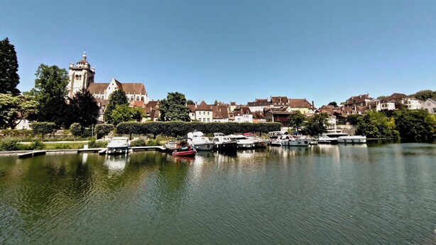 A calm river with boats docked, surrounded by historic buildings and a prominent tower. - La Faran'dole - The cosy cocoon on the edge of the greenway. (Dole)