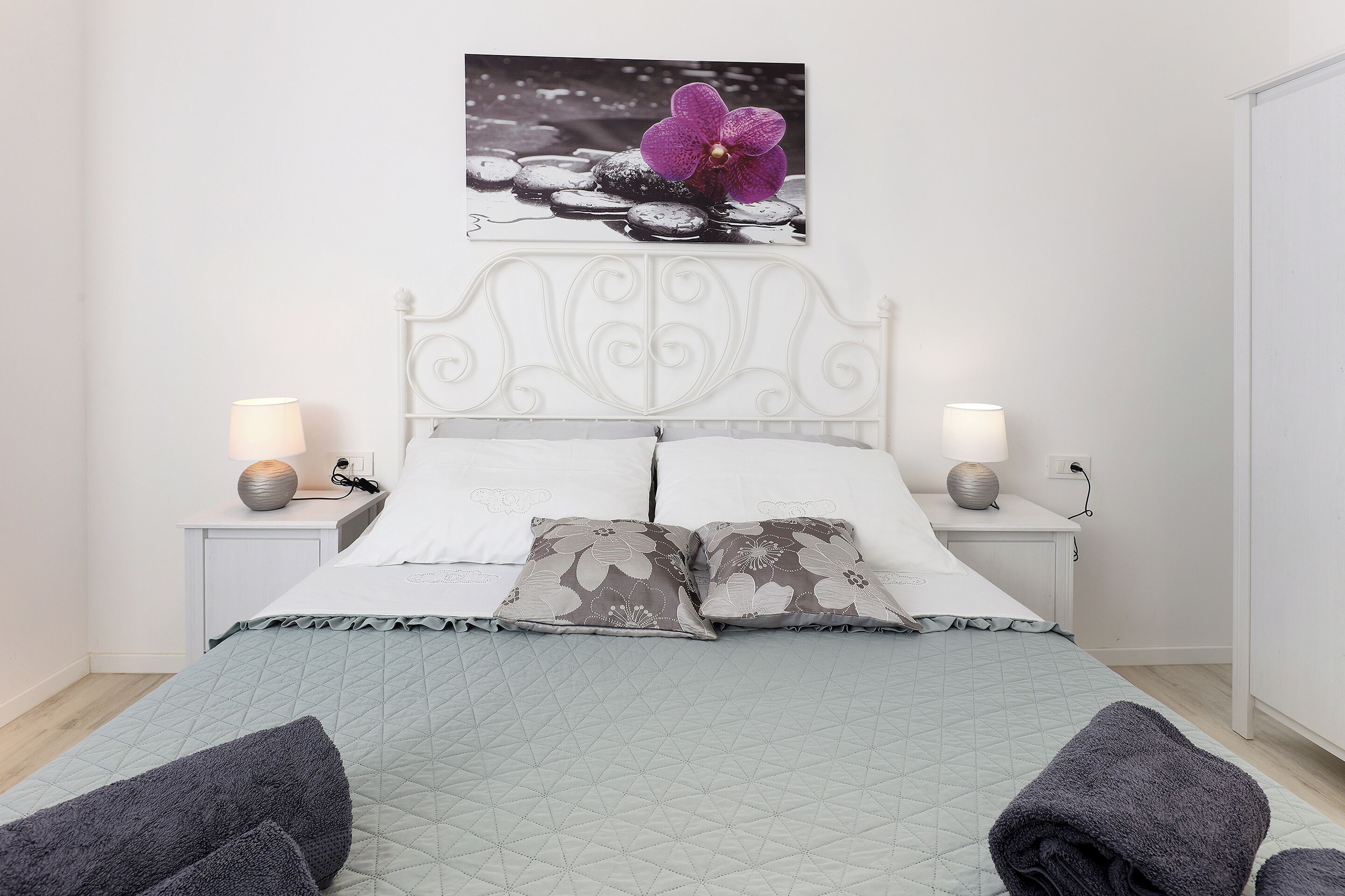 A neatly made bed with a quilted cover, two bedside lamps, and a wall-mounted picture of a purple orchid.