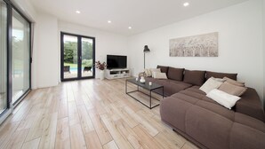 Ein modernes Wohnzimmer mit einem großen braunen Sofa, einem Glastisch und einem Flachbildfernseher. Ein großes Schiebeglasfenster führt auf einen Balkon mit Blick auf Grünflächen.
