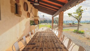 A rustic wooden table with chairs under a covered patio, a brick wall with a window, and a view of a green landscape. - Can Feu: Casa rural, 16-18 personas en Sant Antoni de Vilamajor, 30' Barcelona (Sant Antoni de Vilamajor)