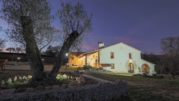 A two-story house with a stone foundation, a large tree in the front yard, and a well-lit entrance. - Can Feu: Casa rural, 16-18 personas en Sant Antoni de Vilamajor, 30' Barcelona (Sant Antoni de Vilamajor)