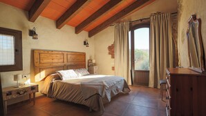 A bedroom with a wooden headboard, a bed with a beige bedspread, a nightstand with a lamp, a dresser, and a window with curtains. - Can Feu: Casa rural, 16-18 personas en Sant Antoni de Vilamajor, 30' Barcelona (Sant Antoni de Vilamajor)