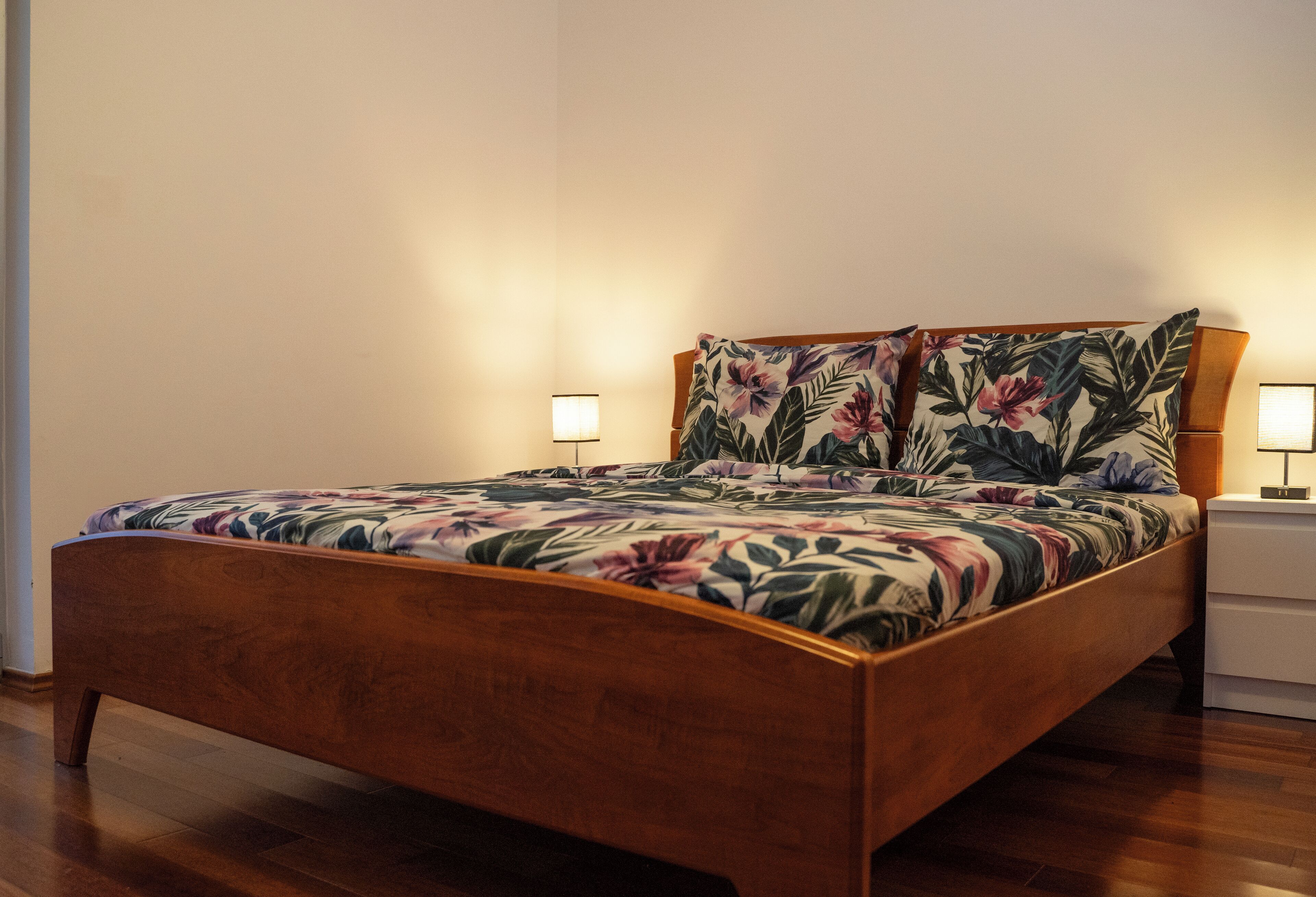 Un lit en bois, recouvert d’une couvre-lit à motifs floraux et agrémenté d’oreillers assortis, avec deux tables de chevet de chaque côté, sur lesquelles sont posées des lampes.