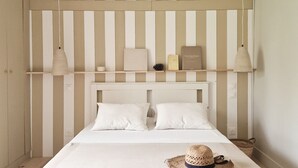 Ein Bett mit weißem Kopfteil, zwei Kissen, einer weißen Decke und einem Strohhut darauf. Zwei Nachttischlampen und ein Buch befinden sich auf dem Kopfteil.