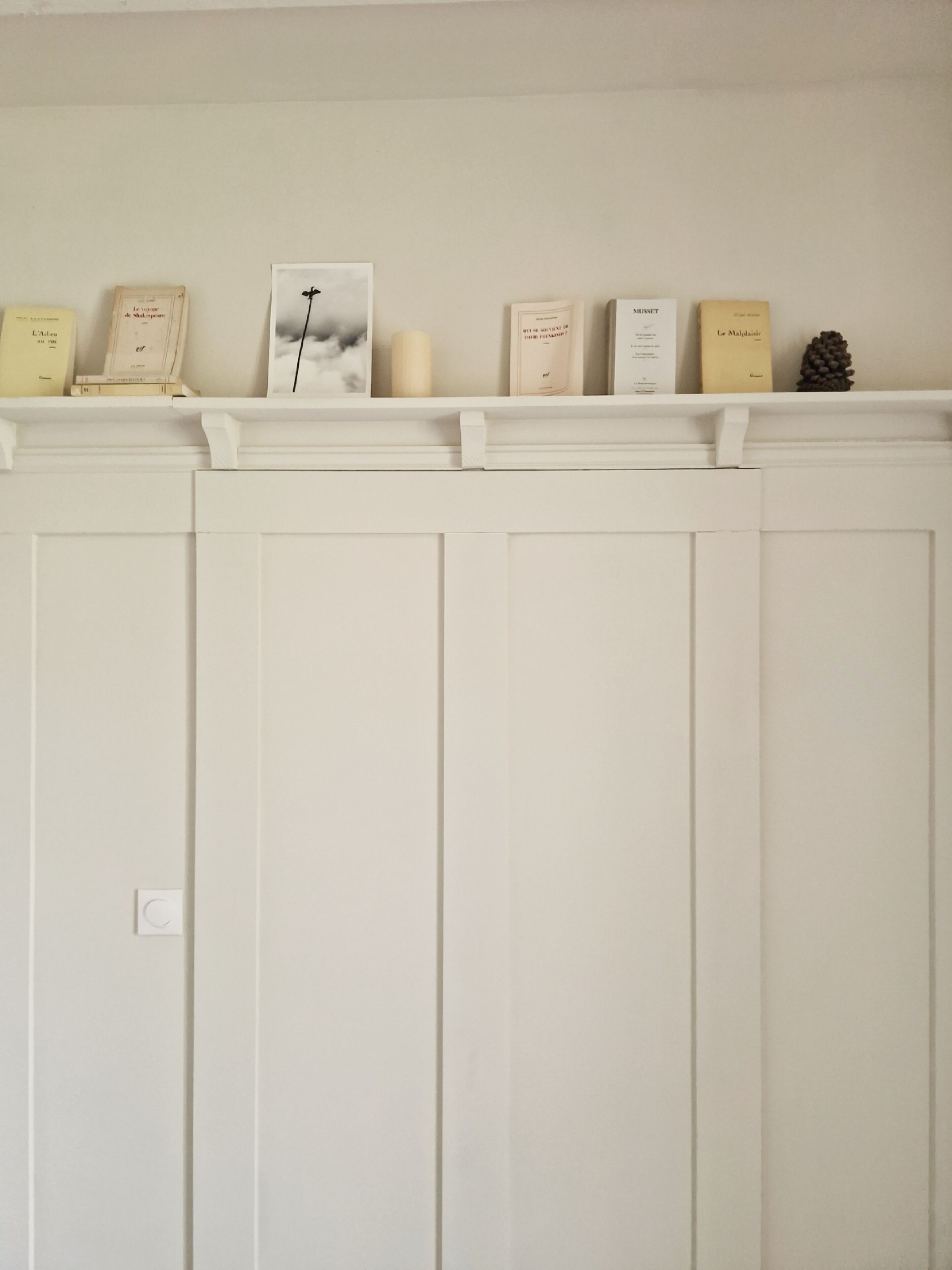 Pared blanca con una repisa que sostiene libros y velas.