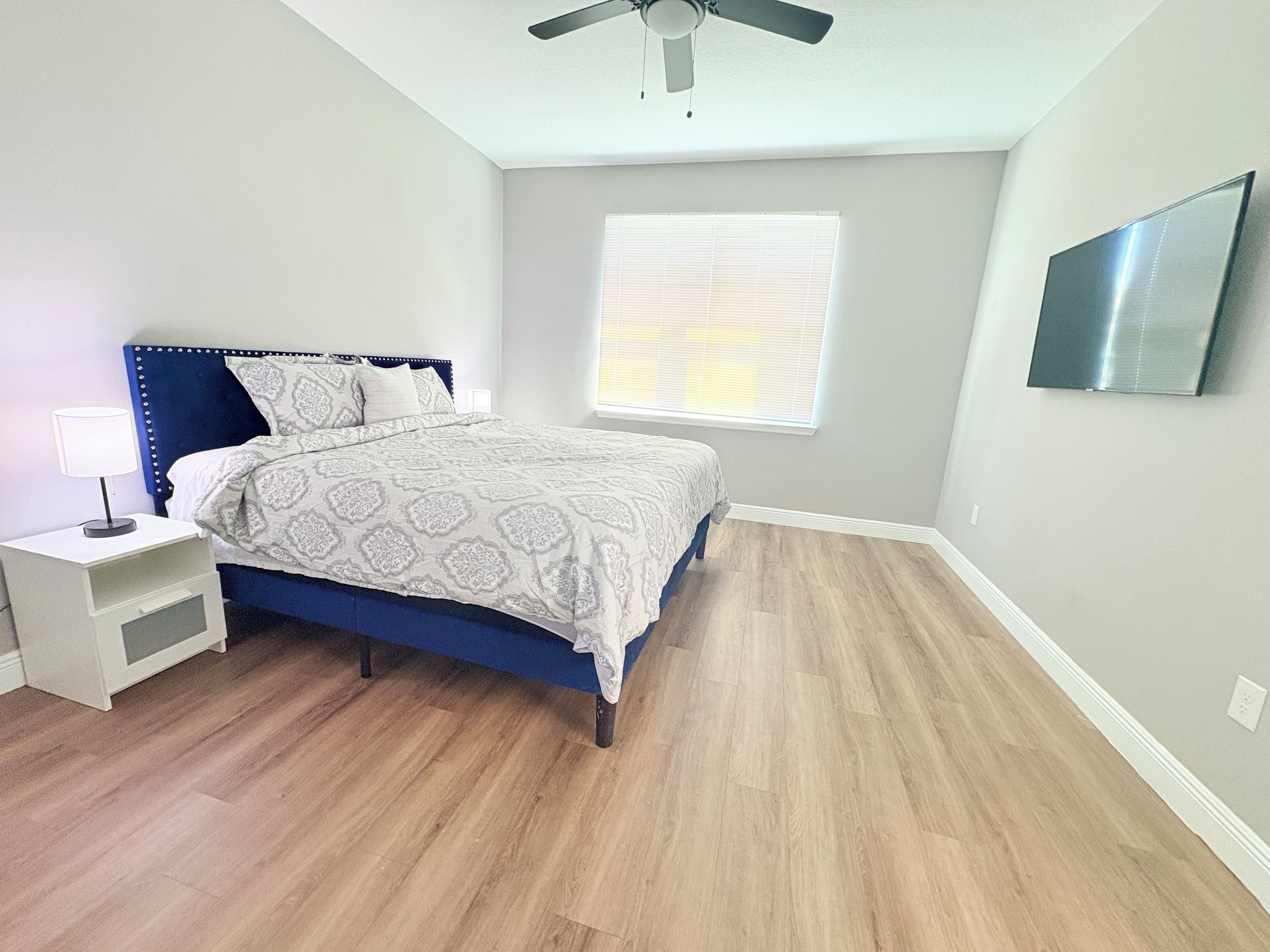 Une chambre à coucher comprenant un lit, une table de chevet, un ventilateur de plafond, une fenêtre et un téléviseur fixé au mur.