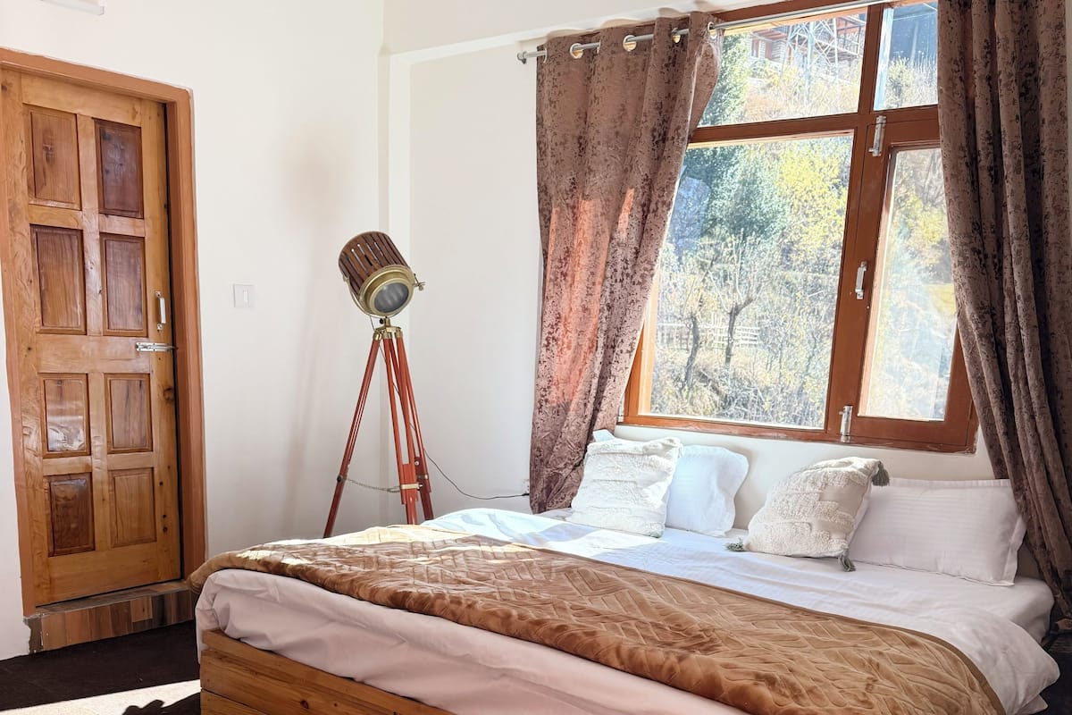 Ein hölzernes Bett mit einer beigen und weißen Bettdecke, ein Holzboden, ein weißer Teppich, eine Holztür und ein Fenster mit Vorhängen.