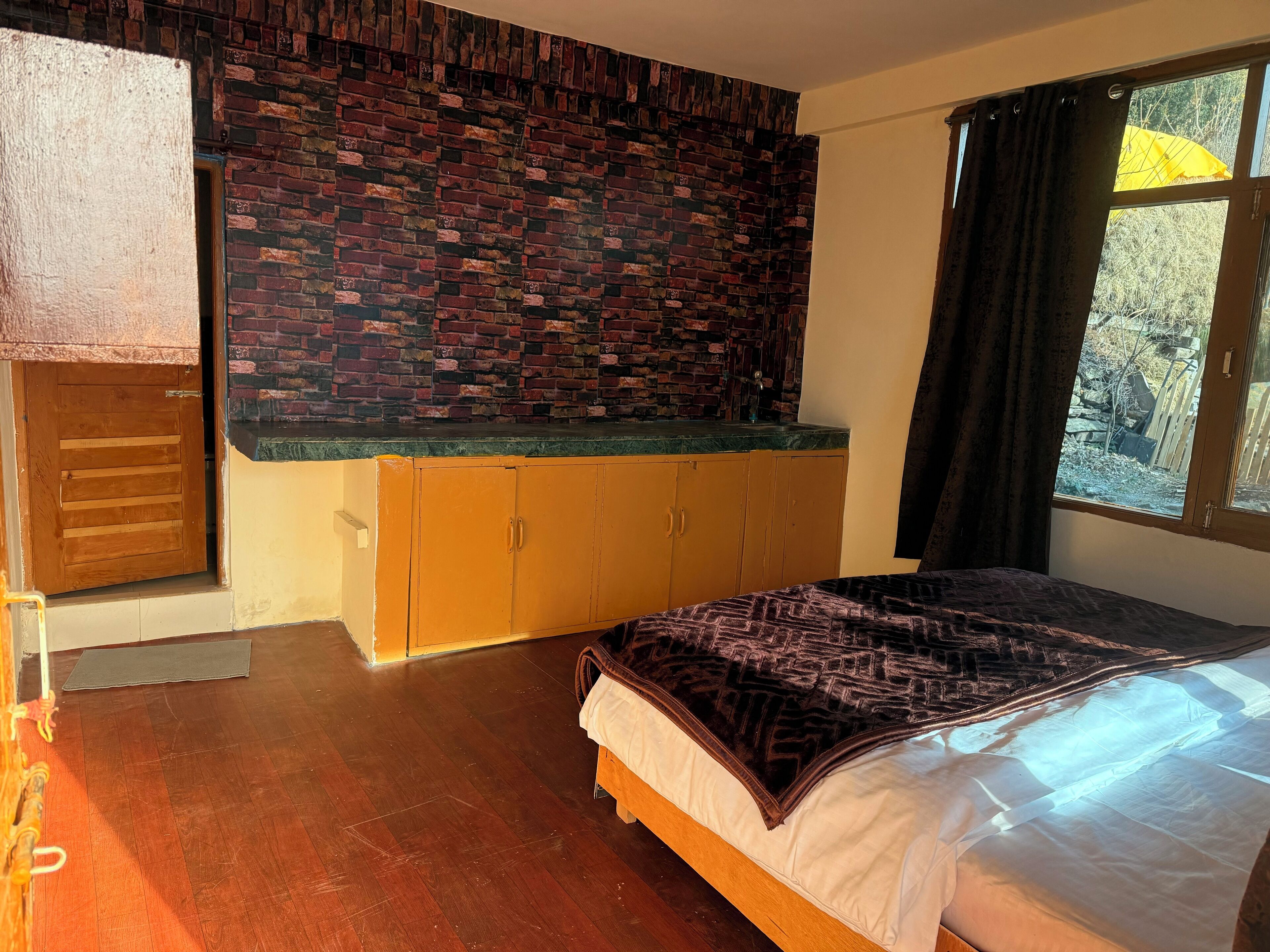 A bedroom with a brick wall, wooden wardrobe, and a bed with a patterned blanket.