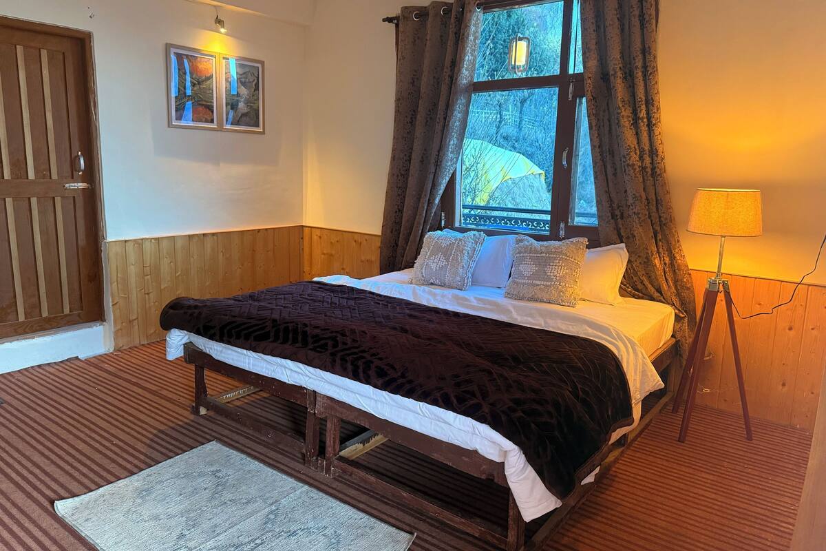 Ein Bett mit Holzrahmen, eine dunkle, gemusterte Bettdecke, weiße Kissen und ein heller Teppich in einem Zimmer mit Holzwänden, einem Fenster mit Vorhängen und einer an der Wand befestigten Lampe.
