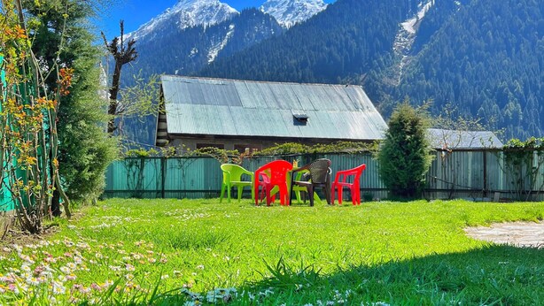 A rural setting with a green field, colorful plastic chairs, a corrugated metal building, and snow-capped mountains in the background. - Kolahoi Heights  (Pahalgam)