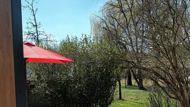 Een terras met een tafel en stoelen onder een rode parasol, omringd door grind en groen.