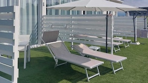 A modern outdoor lounge area with white sun loungers and umbrellas, adjacent to a building with large windows and a white exterior. - Luxury pool Suites - Città Bianca (Spoltore)