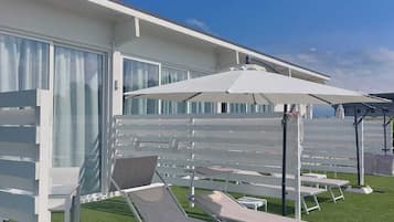 A modern outdoor lounge area with white sun loungers and umbrellas, adjacent to a building with large windows and a white exterior.