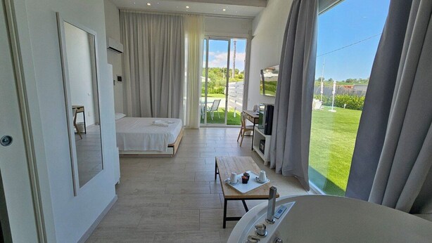 Habitación de hotel con cama, escritorio, silla, mesa con tazas y un mando a distancia, y un ventanal con cortinas.