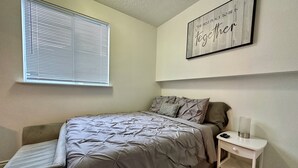 Un dormitorio con cama, mesita de noche y persianas en la ventana. Hay una cita motivacional en la pared.