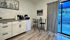Una cocina compacta equipada con microondas, cafetera y un refrigerador pequeño. Hay una mesa de comedor con sillas y una ventana con cortinas.