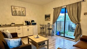 Una sala de estar con sofá, una mesa de centro, una mesa de comedor con sillas, una zona de cocina con armarios y un amplio ventanal corredero que da a un espacio exterior.