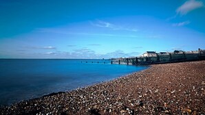 A pebbled beach with a pier extending into the sea, a clear sky, and buildings in the background. - NEW Cossington House - City Centre 3 Bedroom Home (Kent)
