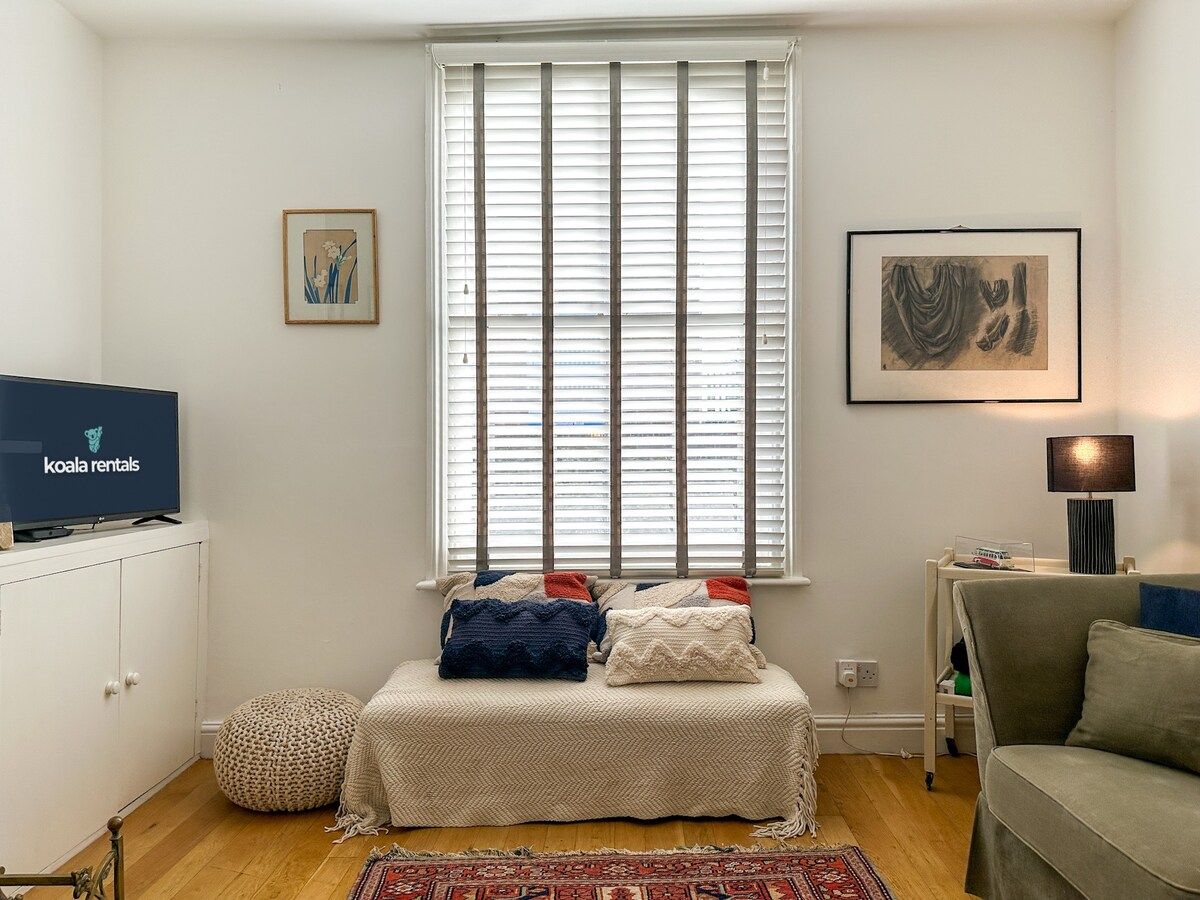 A small, cozy room with a bed, a television, a window with blinds, a lamp, and a rug.