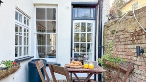 A patio with a wooden table and chairs, a brick wall, and a window with a view of the interior. - NEW Cossington House - City Centre 3 Bedroom Home (Kent)