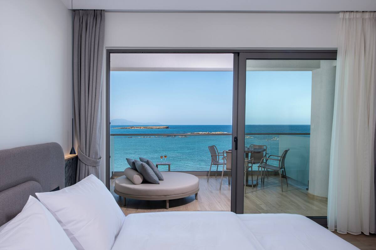 A modern hotel room with a large bed, a seating area with a view of the sea, and a balcony with outdoor seating.