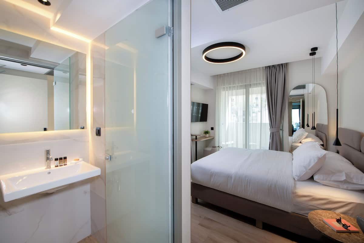 A modern hotel room with a glass shower enclosure, a white sink with toiletries, a large bed with white linens, and a bedside table with a book.