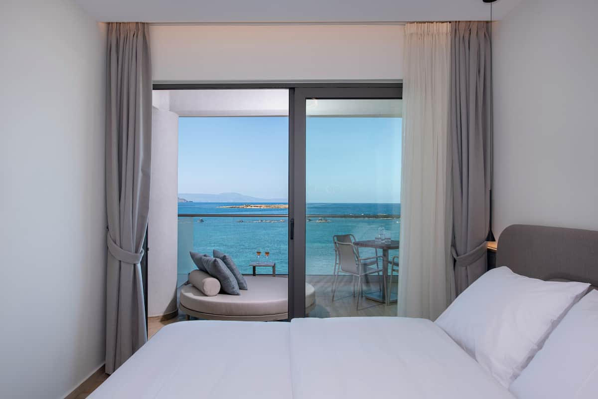 A hotel room with a large bed, a small seating area with a table, and a balcony with a view of the sea.