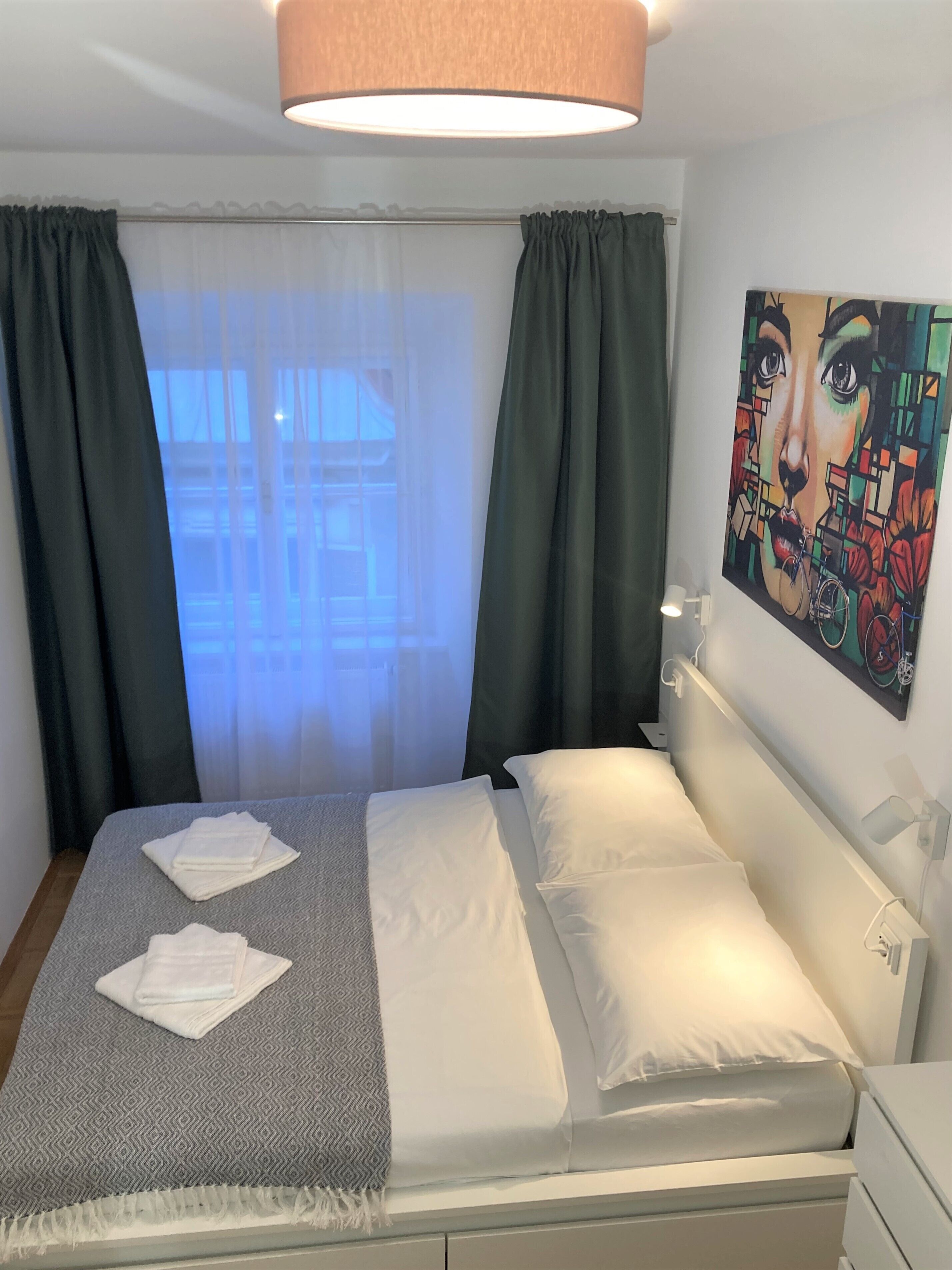 Ett hotellrum med en säng, en tavla på väggen och ett fönster med gardiner.