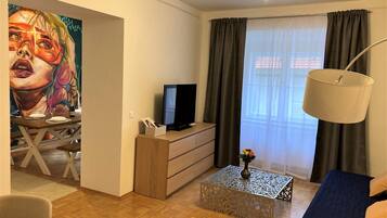 A modern living room with a wooden floor, a sofa, a coffee table, a TV, a painting on the wall, and a window with curtains.